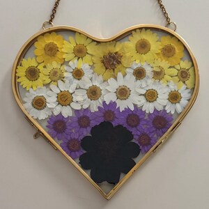 May include: Heart-shaped pressed flower art in a gold frame. The arrangement features yellow, white, purple, and black flowers, creating a non-binary pride flag design. A delicate gold chain is attached for hanging.
