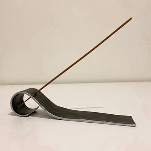 Puede incluir: Un porta incienso de metal con un diseño curvo. Un palo de incienso marrón se coloca en el porta incienso.