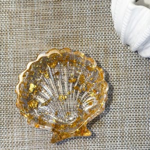 Pode incluir: Um prato transparente em forma de concha com detalhes em folha de ouro. O prato tem uma borda ondulada e um design em forma de leque. A folha de ouro é espalhada pela resina transparente, criando um efeito brilhante. O prato está sobre uma superfície neutra.