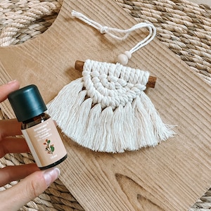 Puede incluir: Un colgante de pared de macramé hecho a mano con una varilla de madera y flecos. Se sostiene en la mano un pequeño frasco de aceite esencial con tapa verde y etiqueta que dice "Sándalo". El fondo es una tabla de cortar de madera.