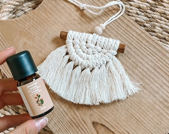 May include: A handmade macrame wall hanging with a natural wood dowel and fringe. A small bottle of essential oil with a green cap and a label that reads "Sandalwood" is held in the hand. The background is a wooden cutting board.
