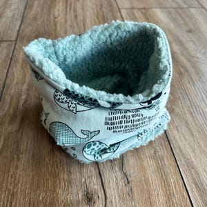 Könnte beinhalten: Ein hellblau-weißes Nackenwärmer mit Walmuster und einem weichen, flauschigen, hellblauen Innenfutter. Der Nackenwärmer ist aus einem weichen Stoff gefertigt und soll den Hals warm halten. Das Walmuster enthält verschiedene Walillustrationen.