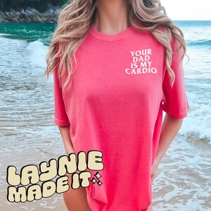 May include: A pink oversized t-shirt with the text "YOUR DAD IS MY CARDIO" printed on the front. The shirt is worn by a person standing on a beach.
