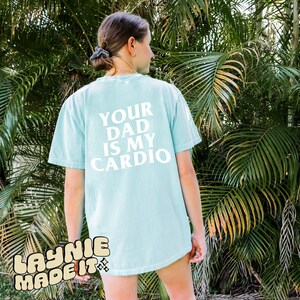 May include: A light blue t-shirt with the text "YOUR DAD IS MY CARDIO" printed in white on the back. The shirt has a distressed look and is made by "LAYNIE MADE IT".