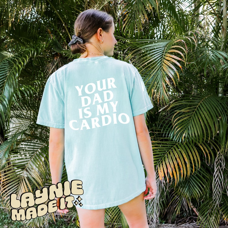 May include: A light blue t-shirt with the text "YOUR DAD IS MY CARDIO" printed in white on the back. The shirt has a distressed look and is made by "LAYNIE MADE IT".