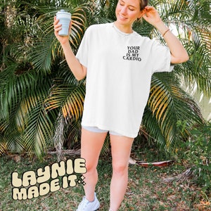 May include: White t-shirt with the text "Your Dad Is My Cardio" printed on the front. The shirt is being worn by a person standing in front of a green leafy background.