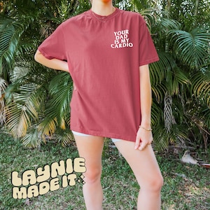 May include: A red t-shirt with the text "Your Dad Is My Cardio" printed in white on the front. The shirt is being worn by a person standing in front of green foliage.