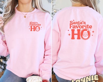 May include: Pink sweatshirt with the text "Santa's Favorite Ho" in red with a Santa Claus graphic on the front and back. The sweatshirt is worn by a person sitting on a wooden stool. The text "Laynie Made It" is on the stool.