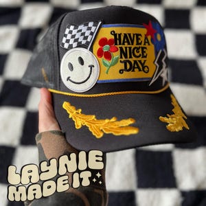 May include: Black trucker hat with a variety of colorful patches, including a smiley face, a checkered flag, a flower, and a patch that says "Have a Nice Day".