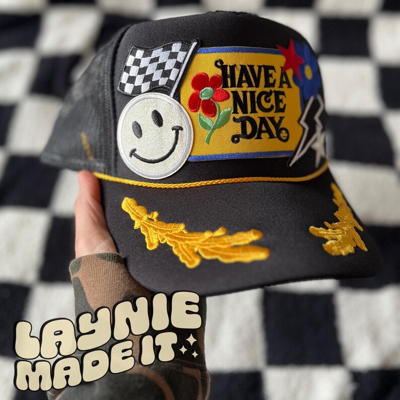 May include: Black trucker hat with a variety of colorful patches, including a smiley face, a checkered flag, a flower, and a patch that says "Have a Nice Day".