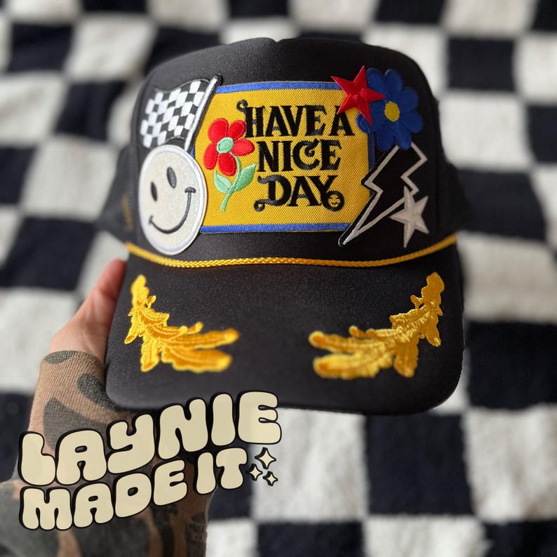 May include: Black trucker hat with a yellow brim and various colorful patches. The patches include a smiley face, a checkered flag, a flower, a lightning bolt, a star, and a patch that says "Have a Nice Day".