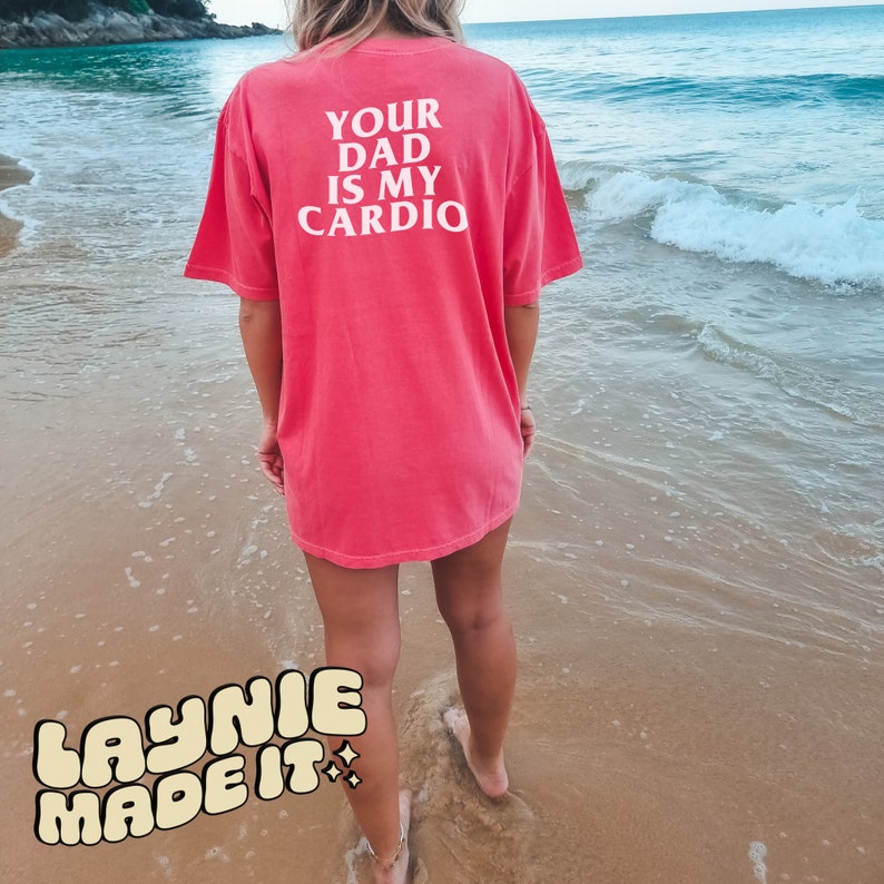 May include: A pink t-shirt with the text "Your Dad Is My Cardio" printed on the back. The shirt is worn by a person standing on a sandy beach.
