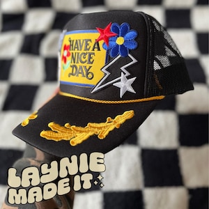 May include: Black trucker hat with a yellow and blue patch that says "Have a Nice Day". The hat also has a white star, a lightning bolt, and a flower patch.