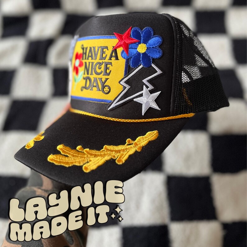 May include: Black trucker hat with a yellow and blue patch that says "Have a Nice Day". The hat also has a white star, a lightning bolt, and a flower patch.