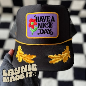 May include: Black trucker hat with a yellow brim and gold embroidered leaves. The front of the hat features a purple patch with a red flower and the text "HAVE A NICE DAY".