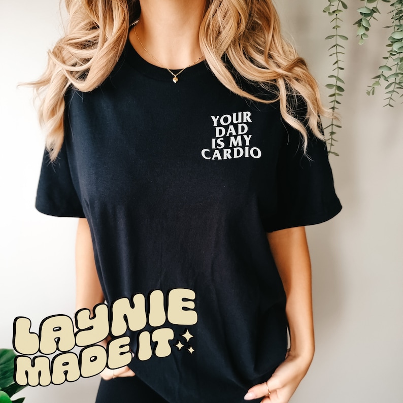May include: Black t-shirt with white text that reads "Your Dad Is My Cardio". The shirt has a yellow and white logo that reads "Laynie Made It" on the bottom left corner.