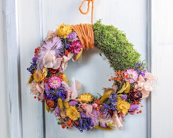 Può includere: Una ghirlanda colorata fatta di fiori secchi, muschio e ramoscelli. La ghirlanda è decorata con fiori viola, rosa, gialli e arancioni. È appesa a una porta di legno bianco.