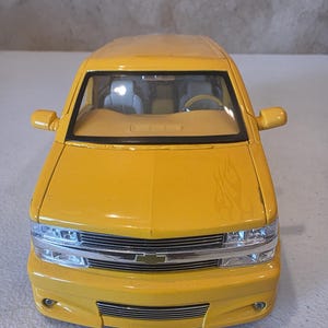 May include: A bright yellow toy truck with a detailed front grille, headlights, and side mirrors. The miniature vehicle has a flame design on the side and a tan interior. The toy truck is a model of a pickup truck.