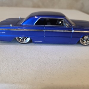 May include: A diecast model of a blue classic car with chrome wheels. The car has a sleek design, with the word "IMPALA" visible on the side. The text "OLD SCHOOL" is also visible. The car is a coupe.