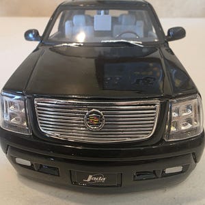 Puede incluir: Un coche de juguete Cadillac Escalade negro con un acabado brillante. La parte delantera presenta una parrilla cromada con el emblema de Cadillac y faros transparentes. El coche de juguete tiene el logotipo "Jada" en el parachoques delantero.