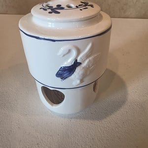 May include: White ceramic container with a lid, featuring blue accents and a swan design. The lid has a blue knob and floral patterns. The container has a heart-shaped opening and is likely for decorative or functional use.