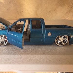 May include: A blue toy pickup truck with the bonnet open and the driver's side door open. The truck has chrome rims and a detailed interior. The truck is a model of a custom pickup truck.