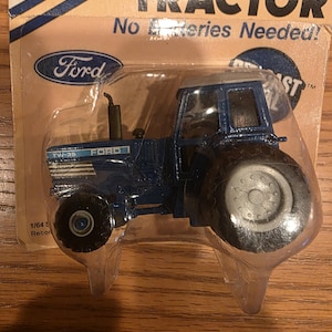 Pode incluir: Um brinquedo de trator Ford TW-35 azul, embalado em plástico transparente. O trator de brinquedo tem pneus pretos e um teto prateado. A embalagem diz "POWER PULL TRACTOR" e "No Batteries Needed!"