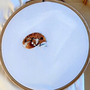 May include: A white embroidery hoop with a brown fox design stitched on it.  There are several spools of embroidery floss in different colors, including white, brown, and orange, with labels that say "Blanc", "976", "310", and "400".