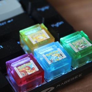 Può includere: Quattro tasti in plastica trasparente con design di cartucce di gioco Pokemon. I tasti sono di diversi colori: giallo, blu, verde e rosso. I tasti sono su una tastiera nera con la parola "MAGEGEE" stampata nell'angolo in basso a destra.