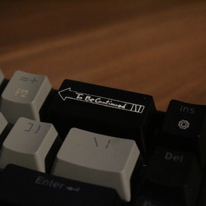Könnte beinhalten: Eine schwarze Tastatur mit einer benutzerdefinierten Taste, auf der "To Be Continued" mit einem weißen Pfeil nach rechts steht. Die Taste ist von anderen schwarzen Tasten mit weißen Beschriftungen umgeben.