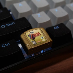 Könnte beinhalten: Eine goldene Nintendo 64-Spielkassette mit dem Text "The Legend of Zelda: Ocarina of Time" auf dem Etikett. Die Kassette liegt auf einer schwarzen Tastatur.