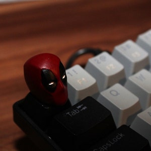 Puede incluir: Un tapón de tecla de teclado mecánico con el personaje Deadpool en rojo y negro. El tapón está colocado en la tecla "Tab" de un teclado blanco y negro.