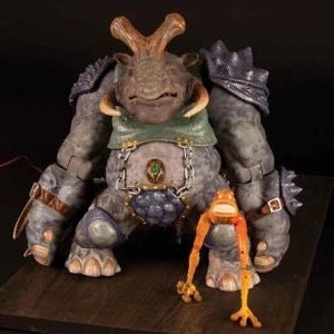 May include: A gray, armored rhinoceros figurine with a green and brown chest piece and a chain. A small orange frog figurine with long legs stands in front of the rhinoceros.