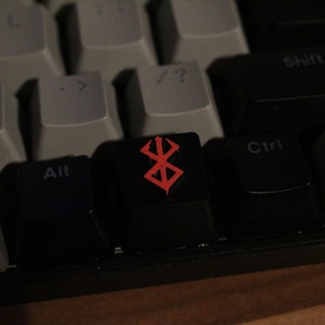 Könnte beinhalten: Nahaufnahme einer mechanischen Tastatur mit grauen und schwarzen Tasten. Die schwarze Taste in der Mitte zeigt ein rotes Symbol. Die Taste ist von anderen Tasten mit weißer Beschriftung umgeben. Die Tastatur befindet sich auf einer Holzoberfläche.