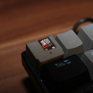 May include: A close-up of a gray and black keyboard with a small gray cartridge labeled "Super Mario Bros." inserted into the top right corner of the keyboard.