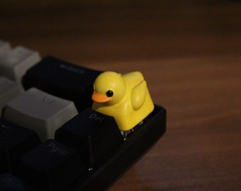 May include: A yellow rubber ducky keycap sits on a black keyboard. The ducky has a small orange beak and black eyes. The keyboard has keys labeled "Del" and "PgUp".