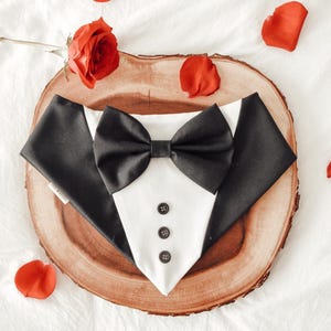 May include: A black and white tuxedo-style dog bandana with a black bow tie and four black buttons. The bandana is on a wooden surface with red rose petals scattered around it.
