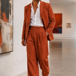 Puede incluir: Un traje de color óxido compuesto por una americana de botonadura sencilla con solapas de muesca y pantalones anchos a juego con pliegues frontales, combinado con una camisa blanca y zapatos de vestir de cuero marrón oscuro.