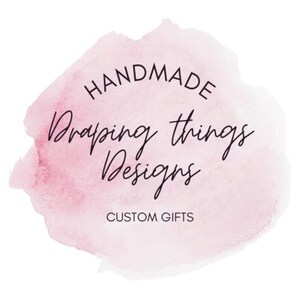 May include: A circular logo design featuring a soft pink watercolor wash background. The text reads HANDMADE in a simple sans-serif font, followed by the script text Draping things Designs, and the words CUSTOM GIFTS in a clean sans-serif font.