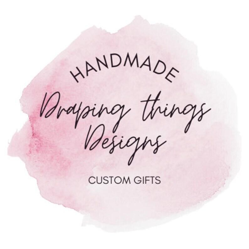 May include: A circular logo design featuring a soft pink watercolor wash background. The text reads HANDMADE in a simple sans-serif font, followed by the script text Draping things Designs, and the words CUSTOM GIFTS in a clean sans-serif font.