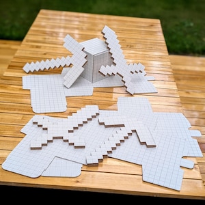 Pode incluir: Uma coleção de recortes de papelão branco com formas pixeladas de espadas e picaretas com um padrão de grade impresso. O conjunto inclui peças planas não montadas e um cubo montado, feitos de material de papelão ondulado.