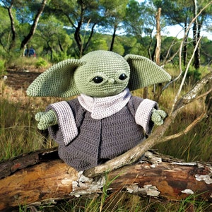 Puede incluir: Un juguete de peluche verde tejido a crochet que se asemeja a un personaje de la franquicia Star Wars, sentado en una rama de árbol en un entorno de bosque. El juguete lleva una túnica gris con capucha y un ribete de color morado claro.