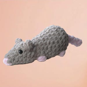 May include: A crocheted rat toy in grey with pink details. The toy features a textured body, small black eyes, and pink nose, tail, and feet. This soft toy is suitable for children and animal enthusiasts.