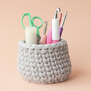 May include: A light gray, crocheted basket filled with craft supplies. The basket contains green scissors, a white candle, and various crochet hooks in pink, purple, and silver. The basket is round and sits on a peach-colored surface.