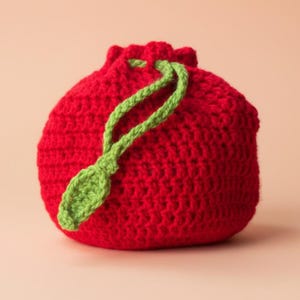 May include: A handmade red crochet drawstring bag with a green leaf accent. The bag is round and has a textured surface. The drawstring and leaf are a bright green color, contrasting with the red body of the bag.