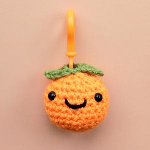 May include: A crocheted orange fruit charm with a smiling face, black eyes, and green leaves. The orange charm has an attached orange plastic clip. The background is a solid peach color.