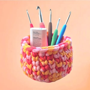May include: A colorful, handmade crochet basket in shades of pink, red, and yellow, holding various crochet hooks. The hooks have different colored handles, including pink, brown, green, and blue. A white cylindrical container with text is also inside the basket.