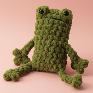 May include: A handmade green crochet frog plush toy with black eyes. The frog has outstretched arms and legs, and is sitting on a pink background. The plush toy is made of a textured yarn.