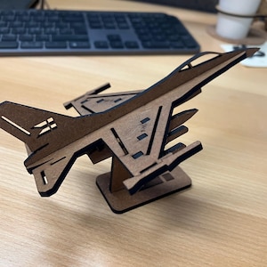 May include: A wooden model of a fighter jet, featuring a brown body with black accents. The model is displayed on a small stand, showcasing the wings, tail, and cockpit details. The jet is approximately 6 inches long.