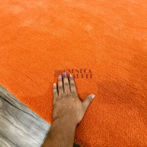 May include: A solid bright orange area rug with a dense, plush, and soft-textured pile. The surface features a small, dark, rectangular logo stamp in the center that reads SENECA CARPET in a clear, sans-serif font, indicating the brand name.